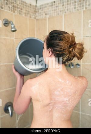 Économisez de l'eau, de l'argent et de la planète. Vue arrière d'une femme méconnue lavant son corps avec un seau d'eau dans la salle de bains à la maison. Banque D'Images