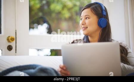 Elle a tout couvert pour le week-end. C'est une jeune femme très attirante qui porte des écouteurs tout en utilisant un ordinateur portable à la maison. Banque D'Images