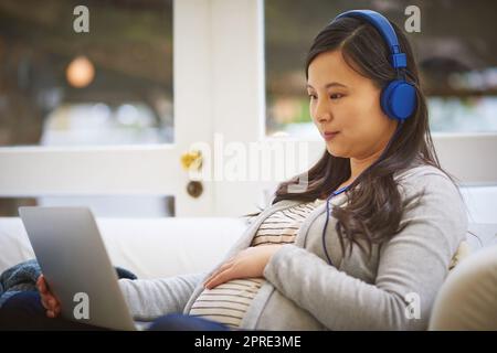 Liée à tout ce qu'elle a besoin de savoir en préparation à la maternité. Une femme enceinte portant un casque tout en utilisant un ordinateur portable à la maison. Banque D'Images