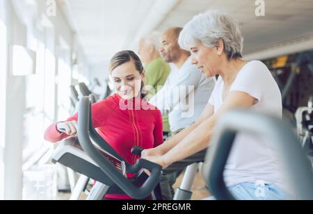 Fitness coach in gym helping senior people during cardio bike training Stock Photo