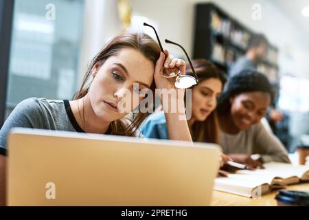 Faire la fête hier soir n'était probablement pas une bonne idée. Une jeune femme utilisant un ordinateur portable dans une bibliothèque d'université et regardant stressée. Banque D'Images