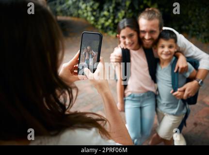 Mère prenant une photo de famille sur son téléphone de ses petits enfants heureux et de leur père souriant avant l'école. Garçon et fille frères et sœurs avec leur père sourire pour une photo. Maman prenant des photos d'enfants Banque D'Images