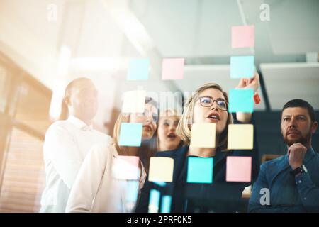 Planifiez votre réussite et soyez prêt à l'obtenir. Un groupe d'hommes d'affaires qui organisent des notes adhésives sur un mur de verre dans un bureau moderne. Banque D'Images