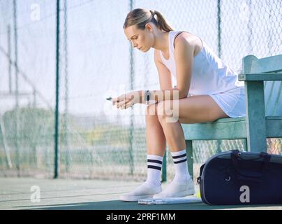 Une joueuse de tennis féminine avec un téléphone vérifiant la progression de l'objectif de fitness sur l'application d'exercice moderne en ligne tout en prenant une pause sur le court. Une femme de sport regardant des messages sur le téléphone portable et attendant l'entraîneur Banque D'Images