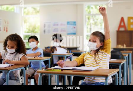 Porter un masque facial pour se protéger des covid pendant l'apprentissage en classe, répondre à une question d'éducation et étudier avec les élèves dans une salle de classe. Fille assise à un bureau et levant la main pendant une pandémie Banque D'Images