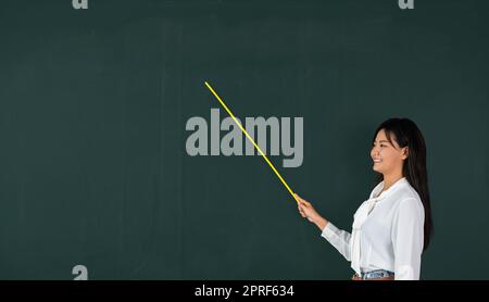 Femme enseignante asiatique souriante avec un bâton de bois pointant vers le tableau noir Banque D'Images