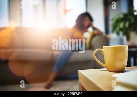 Le café et la détente vont de pair. Gros plan d'une tasse sur une table basse avec une jeune femme méconnue se relaxant en arrière-plan. Banque D'Images