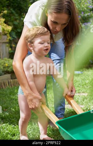 Apprendre le jardinage. Un bébé garçon poussant sa roue de jouet barrow pendant que sa mère aide Banque D'Images