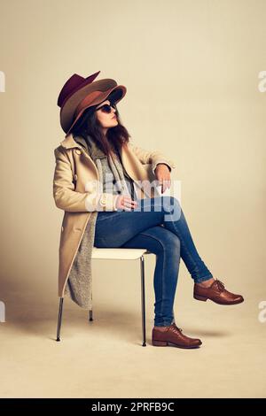 Découvrez les dernières tendances en matière de chapeaux. Photo studio d'une jeune femme assise sur une chaise et portant un tas de chapeaux sur fond marron. Banque D'Images