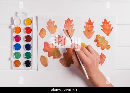 Femelle dessin à la main de feuilles d'automne de différentes formes, tailles et couleurs avec aquarelles et pinceau sur table blanche en bois. Vue de dessus. Automobile maison à la main Banque D'Images