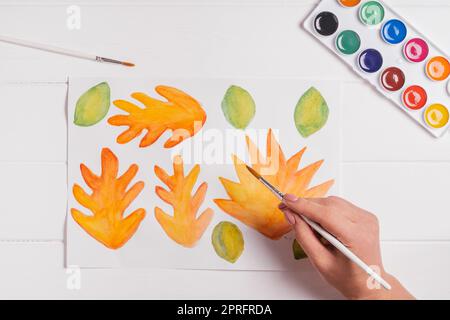 Femelle dessin à la main de feuilles d'automne de différentes formes, tailles et couleurs avec aquarelles et pinceau sur table blanche en bois. Vue de dessus. Automobile maison à la main Banque D'Images