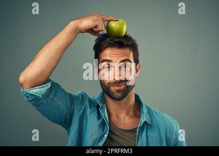 Il s'agit de trouver le bon équilibre. Studio portrait d'un beau jeune homme posant avec une pomme sur sa tête sur un fond gris. Banque D'Images