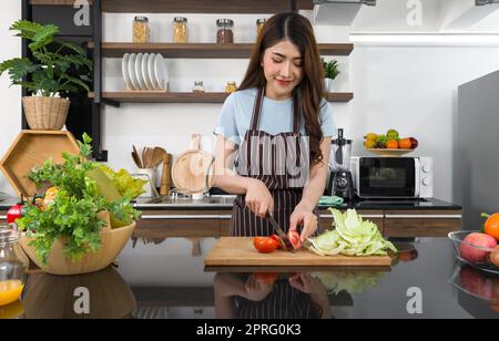 Jeune femme de ménage asiatique vêtue d'un tablier, tranche de tomate et laitue croquante sur une chop en bois. Le comptoir de cuisine plein de différents types de fruits et légumes. Banque D'Images