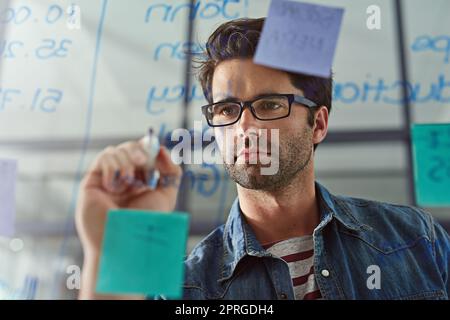 Déterminé à réaliser ses visions de succès. Un jeune designer écrivant des notes sur un mur de verre dans un bureau moderne. Banque D'Images