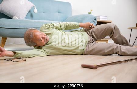 Un vieil homme asiatique allongé sur le sol après être tombé avec un bâton de marche en bois, un homme âgé malade à côté d'un canapé sur un tapis dans la salle de séjour à la maison, ayant des personnes âgées Banque D'Images