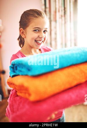 Tout propre et doux. Portrait d'une petite fille gaie portant un tas de serviettes fraîchement lavées tout en regardant l'appareil photo à la maison. Banque D'Images