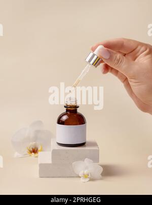 Main avec une pipette sur la bouteille de compte-gouttes cosmétique ouverte près des fleurs d'orchidées blanches, Mockup Banque D'Images