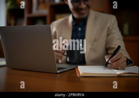 Gros plan d'un homme d'affaires asiatique ou d'un PDG professionnel adulte prenant des notes sur son ordinateur portable et sirotant un café à son bureau dans la salle de bureau. Banque D'Images