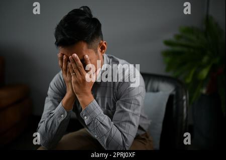 Un homme asiatique millénaire stressé et déprimé couvre son visage, pleurant sur ses échecs dans la vie tout en étant assis dans un salon. Banque D'Images