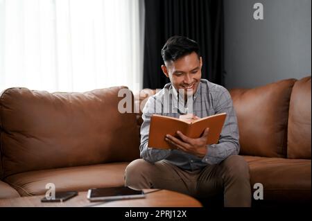 Homme asiatique du millénaire élégant et heureux qui fait la liste ou prend des notes sur son ordinateur portable tout en étant assis sur un canapé dans son salon. Banque D'Images