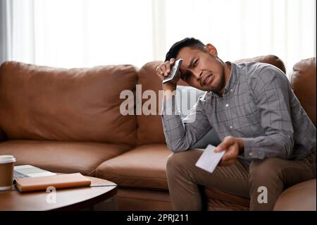 Stressé et malheureux homme asiatique millénaire assis sur un canapé dans un salon se sentant bouleversé et malheureux, ayant un problème financier ou ayant des problèmes avec Banque D'Images