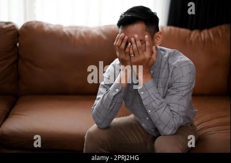 Un homme asiatique millénaire stressé et déprimé couvre son visage, pleurant sur le canapé dans un salon, souffrant de sa vie infructueuse. Banque D'Images