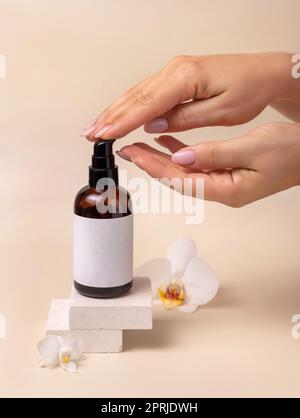 Une femme pompe à la main du liquide sur une main courte d'une bouteille cosmétique près des fleurs d'orchidées sur beige gros plan, maquette d'étiquette. Kit de produits de beauté pour le soin de la peau Banque D'Images
