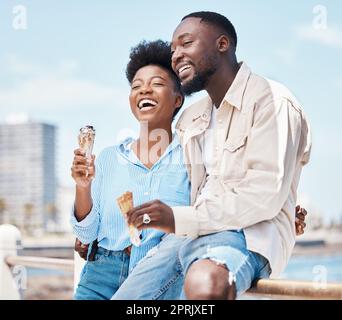 Couple heureux à la plage en train de manger un cône de glace pendant une journée dans la nature pendant les vacances de printemps. Homme noir et femme parlant et riant tout en embrassant et ayant le dessert en vacances en bord de mer. Banque D'Images