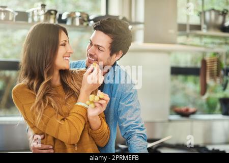 Avoir un raisin. Une jeune femme attrayante nourrissant son petit ami raisins dans leur cuisine. Banque D'Images