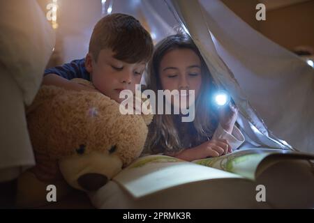 Pourquoi dormir quand vous pourriez lire à la place. Frères et sœurs lisant un livre par torchlight sous une couverture fort à la maison. Banque D'Images