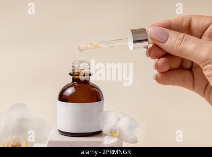 Main avec une pipette sur la bouteille de compte-gouttes cosmétique ouverte près des fleurs d'orchidées blanches, Mockup Banque D'Images