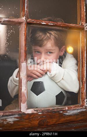 Il pleut à nouveau... Un jeune garçon assis près de la fenêtre et qui avait l'air de s'ennuyer pendant qu'il pleut dehors. Banque D'Images