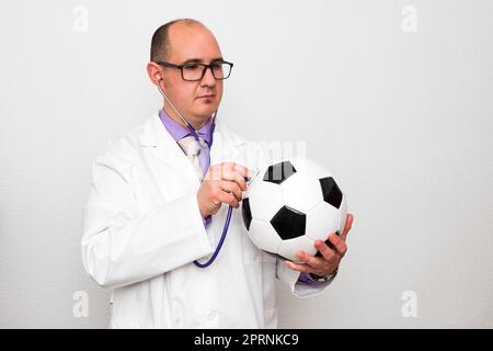 Un médecin caucasien chauve avec des lunettes portant un manteau blanc sur une chemise et une cravate pourpres tient une balle de football tout en l'auscultant avec un stethoscop Banque D'Images