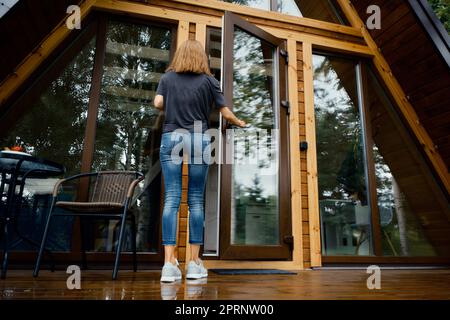 Vue arrière d'une femme ouvrant la porte d'une maison de campagne et entrant à l'intérieur Banque D'Images