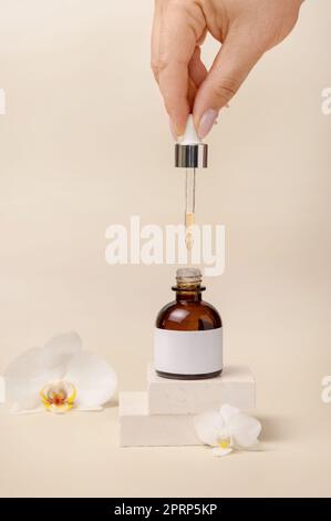 Main avec une pipette sur la bouteille de compte-gouttes cosmétique ouverte près des fleurs d'orchidées blanches, Mockup Banque D'Images