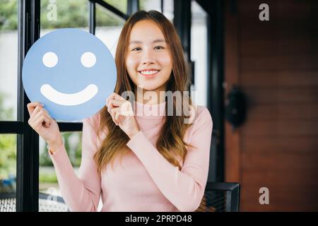 Examen de l'expérience client. Bonne heureuse excitée jeune femme souriante montrant une excellente évaluation avec smiley visage icône sur le papier cercle bleu dans café feedback goo Banque D'Images