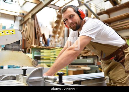 Portrait d'un carpenter en vêtements de travail et de protection de l'ouïe dans l'atelier d'un atelier du menuisier Banque D'Images