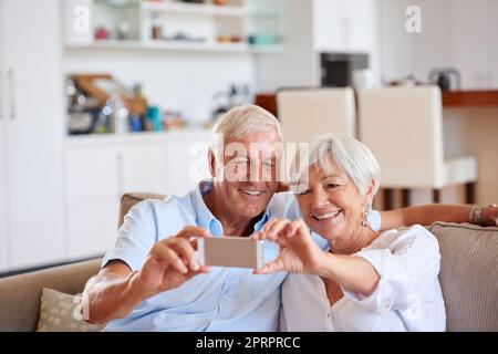 Envoyons celui-ci à nos enfants. Un couple senior prenant un selfie avec son téléphone portable tout en étant assis sur le canapé. Banque D'Images