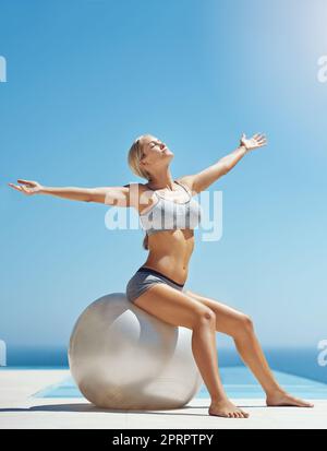 Je n'ai jamais été plus heureux avec mon corps. Prise de vue en longueur d'une jeune femme attirante assise sur une balle d'exercice avec ses bras levés près de la piscine. Banque D'Images