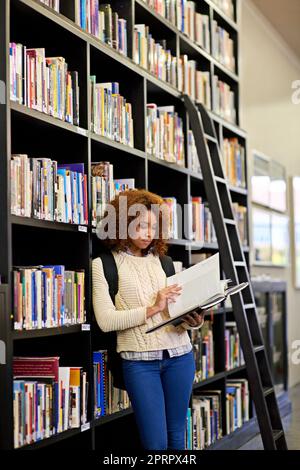 La connaissance est libre à la bibliothèque... il suffit d'apporter un conteneur. Une jeune femme étudie à partir d'un livre tout en se tenant près d'une bibliothèque. Banque D'Images