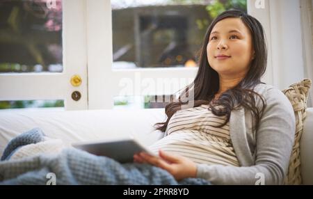 Pas longtemps maintenant jusqu'à ce que le petit arrive. une femme enceinte qui a l'air réfléchie lorsqu'elle utilise une tablette numérique à la maison Banque D'Images