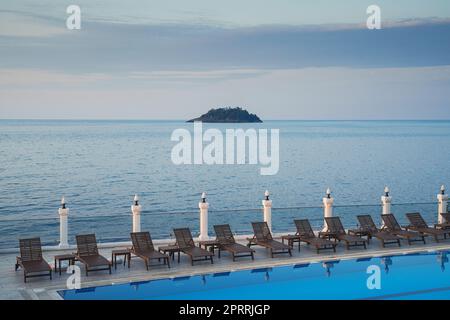 chaises longues en bois près d'une piscine avec mer et une petite île en arrière-plan au coucher du soleil Banque D'Images