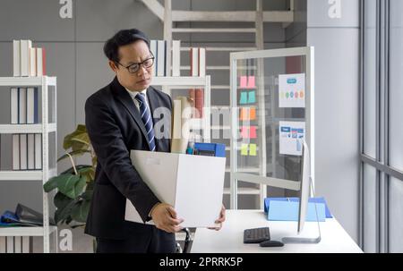 Le Manager asiatique en costume noir se sent triste, stressé et malheureux visage tenant une boîte en papier des effets recueillis au bureau le jour du feu. Idée pour les chômeurs, crise du travail. Banque D'Images