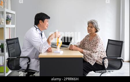 Chirurgien orthopédique asiatique en robe blanche et stéthoscope utilise le modèle d'anatomie des articulations osseuses humaines, présente au patient âgé au sujet de l'arthroplastie totale du genou. Concept de santé et de médecine. Banque D'Images