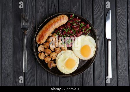 Poêle à frire en fonte avec œufs frits, saucisses, champignons et haricots sur une table en bois. Vue de dessus. Banque D'Images