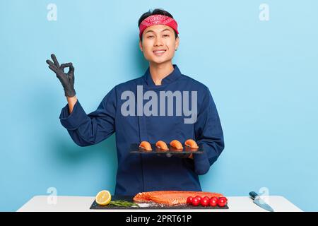 Le jeune homme en blouson bleu du chef se tient près de la table avec du filet de poisson et des tomates, posant avec une assiette de sushis préparés, montre le signe OK avec son rig Banque D'Images