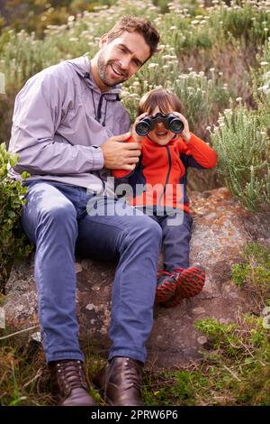 Nous À plus. un petit garçon regardant à travers des jumelles pendant une randonnée avec son père. Banque D'Images