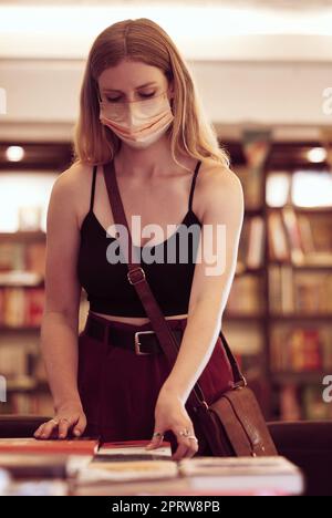 Covid, masque facial et femme avec des livres dans une bibliothèque, une librairie ou un bâtiment éducatif pour la connaissance, la lecture ou la recherche. Jeune étudiante parcourant et choisissant un roman sur table dans une librairie Banque D'Images