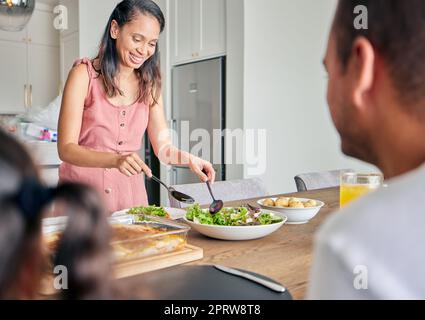 Femme, maman et nourriture saine avec une femme servant le déjeuner ou le dîner pour sa famille avec un sourire à la maison. Bonne femme de ménage préparant une salade verte et dégustant un repas végétalien à la table ensemble Banque D'Images