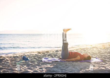 Femme âgée à la plage, sur un tapis de yoga, en train de faire de l'exercice. Lever les jambes. Banque D'Images